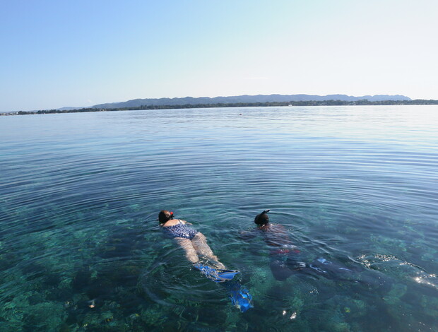 Two people wearing snorkeling gear float face-down in clear, calm water, with a distant shoreline and mountain range visible under a clear blue sky.